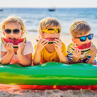 Drei Kinder mit Sonnenbrillen liegen am Strand auf einem aufblasbaren Pool-Float und essen lachend Wassermelone.