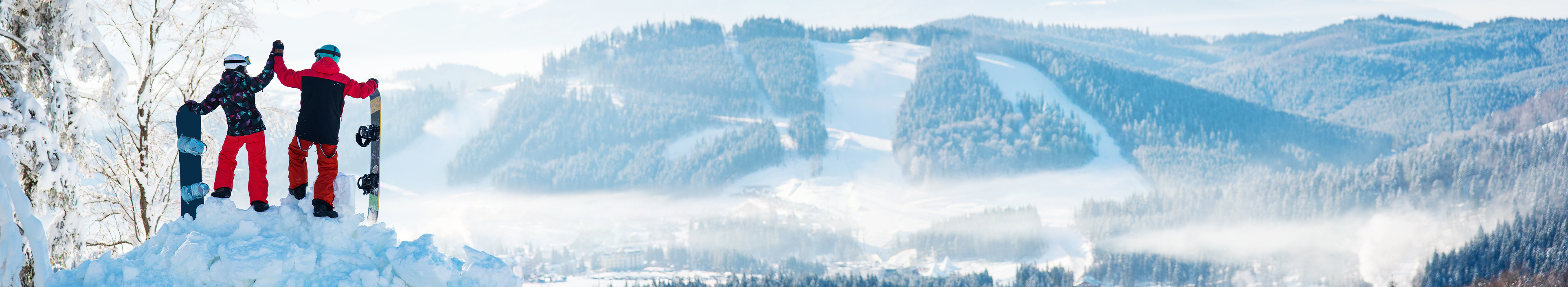 Zwei Snowboarder machen Winterurlaub und stehen auf einem Schneehaufen und halten in der einen Hand ihr Snowboard fest und mit der anderen klatschen sie sich ab. Sie blicken in den verschneiten Tal und die Sonne strahlt.