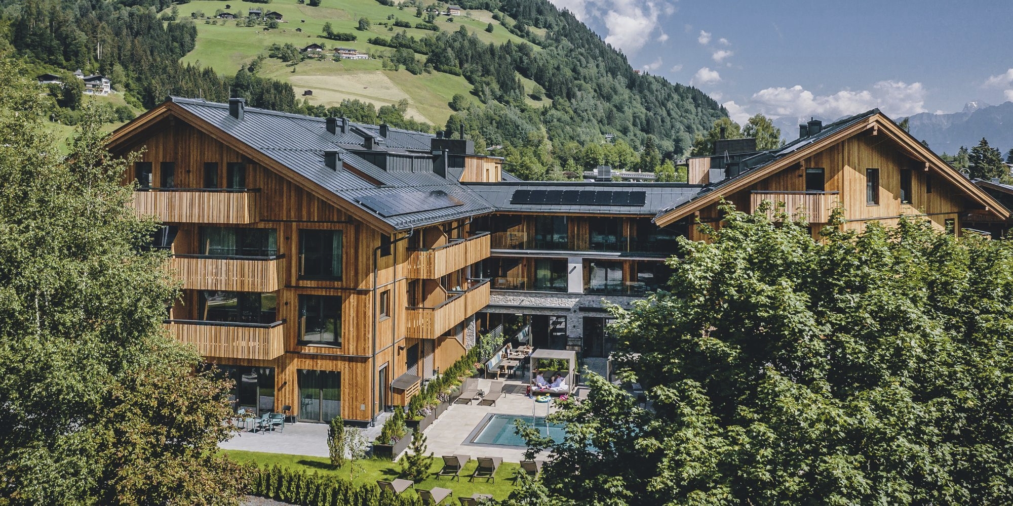 Moderne, U-förmige Hotelanlage im alpinen Chalet-Stil mit hellen Holzfassaden, Balkonen und Solaranlagen auf den Dächern. Im geschützten Innenhof befindet sich eine Terrasse mit einem kleinen Außenpool, umgeben von Sonnenliegen und grünen Rasenflächen vor einem bewaldeten Berghang.