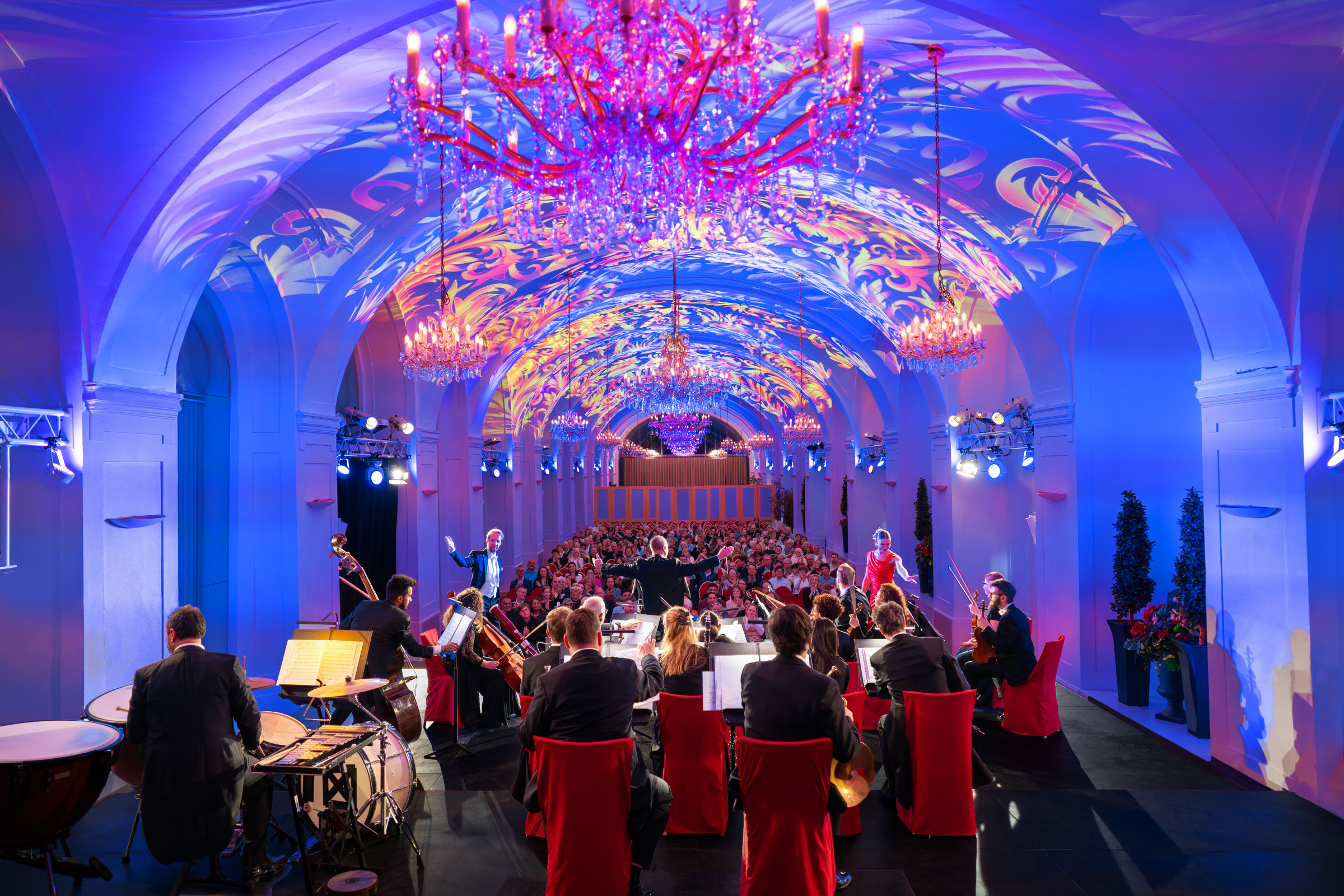 Orchester spielt vor Publikum in einem prunkvoll blau und violett erleuchteten Saal unter Kristallkronleuchtern.