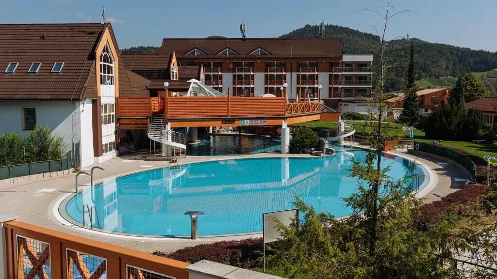 Außenbereich der Terme Zreče mit einem großen Freibad, Holzbrücken und dem Hotel Vital vor bewaldeter Hügelkulisse.