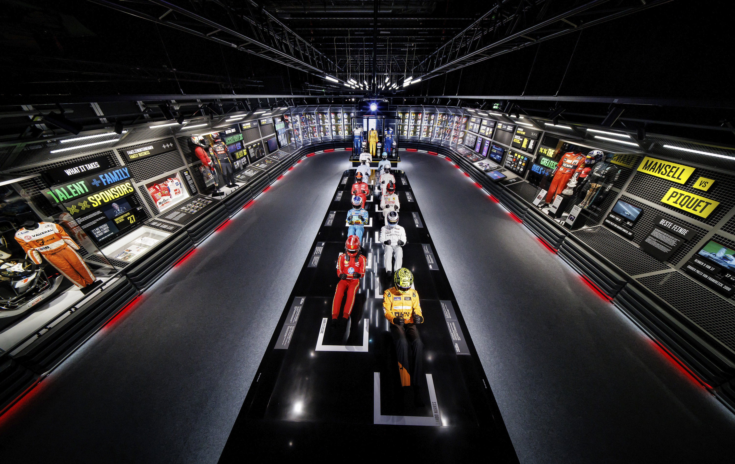 Ein Formel-1-Rennwagen von Max Verstappen (Red Bull Racing) steht in einer dunkel beleuchteten Ausstellungshalle. Im Hintergrund ist ein großes Schwarz-Weiß-Porträt des Fahrers zu sehen. Eine Gruppe junger Besucher betrachtet das Fahrzeug von der Seite. Im Hintergrund läuft ein Video auf einer Leinwand.