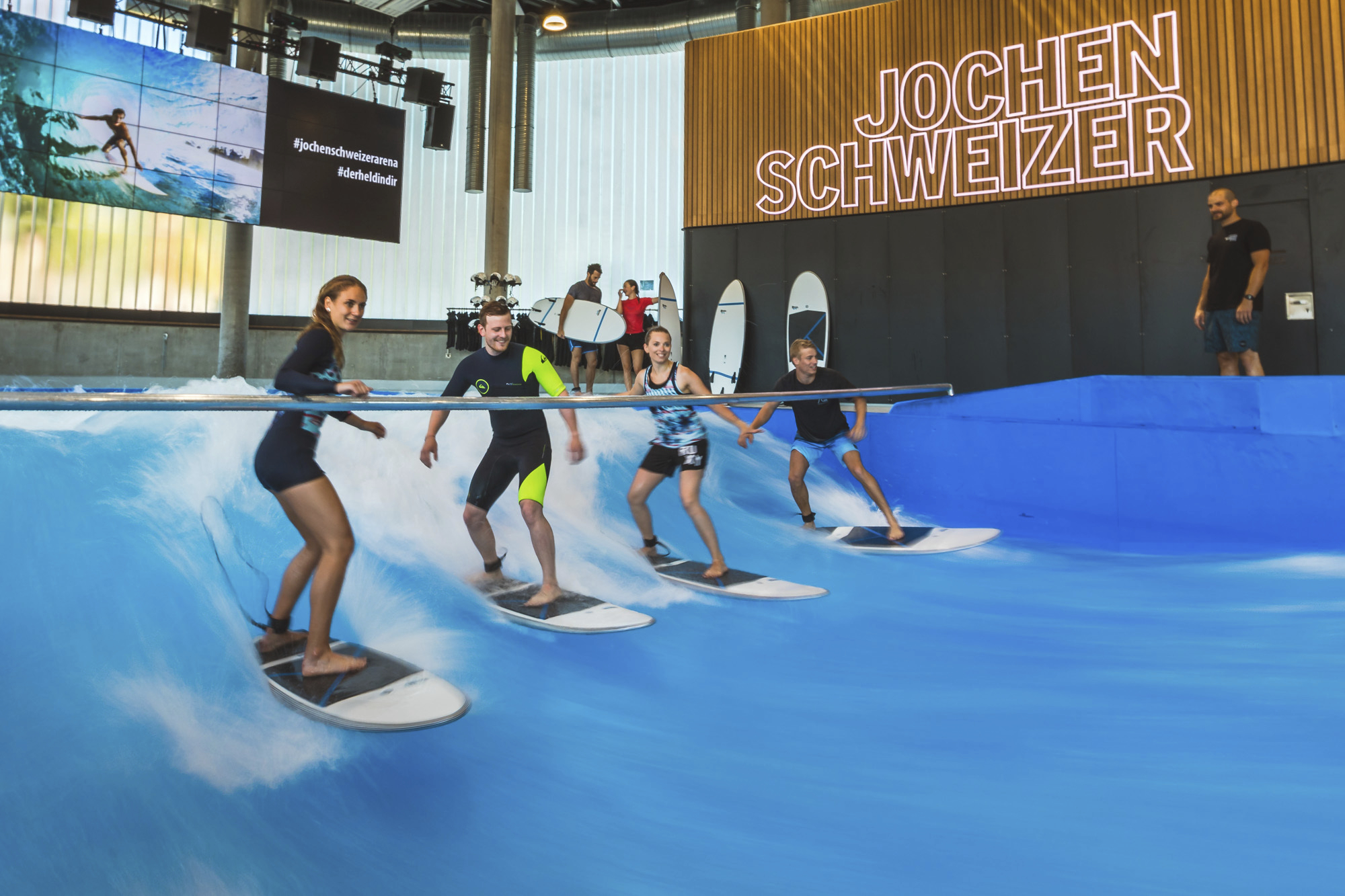 Vier gut gelaunte Surfer (zwei Frauen und zwei Männer) reiten nebeneinander auf einer künstlich erzeugten, stehenden Indoor-Welle. Über dem Becken befindet sich eine Haltestange für Anfänger. Im Hintergrund ist eine Wand mit der großen, leuchtenden Aufschrift „JOCHEN SCHWEIZER“ sowie gelagerten Surfbrettern und Bildschirmen zu sehen.