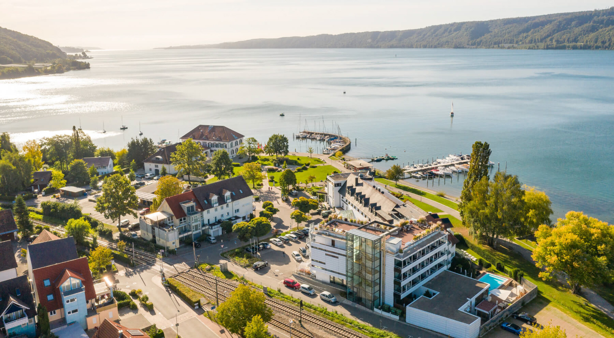 Luftaufnahme einer Hotelanlage am Seeufer mit Bootsstegen, Poolbereich und umliegenden Häusern bei Sonnenschein.
