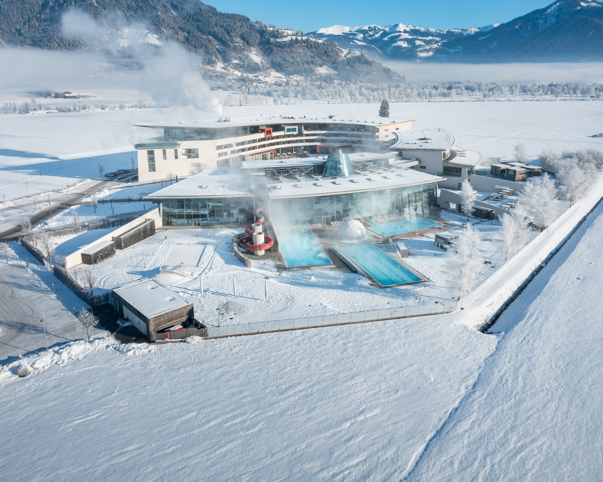 Luftaufnahme der verschneiten Wellnessanlage Tauern Spa Kaprun mit beheizten Außenbecken vor alpiner Kulisse.