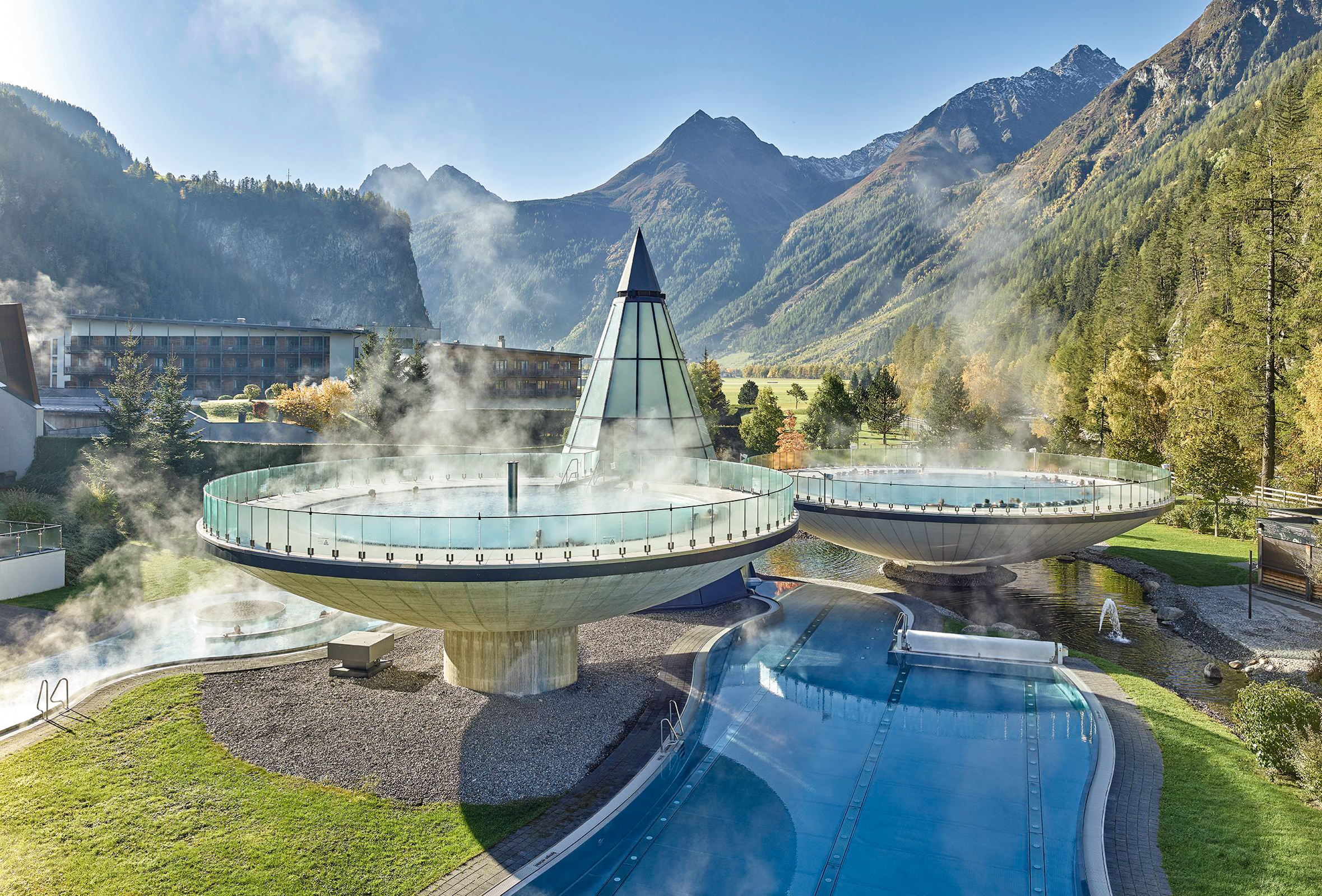 Eine Außenansicht der Therme Aqua Dome in Längenfeld. Im Zentrum stehen zwei riesige, futuristische Schalenbecken, die wie schwebende Kelche über einem Thermalfluss thronen. Dampf steigt vom warmen Wasser in die klare Gebirgsluft auf, während im Hintergrund die imposanten Gipfel der Ötztaler Alpen aufragen.