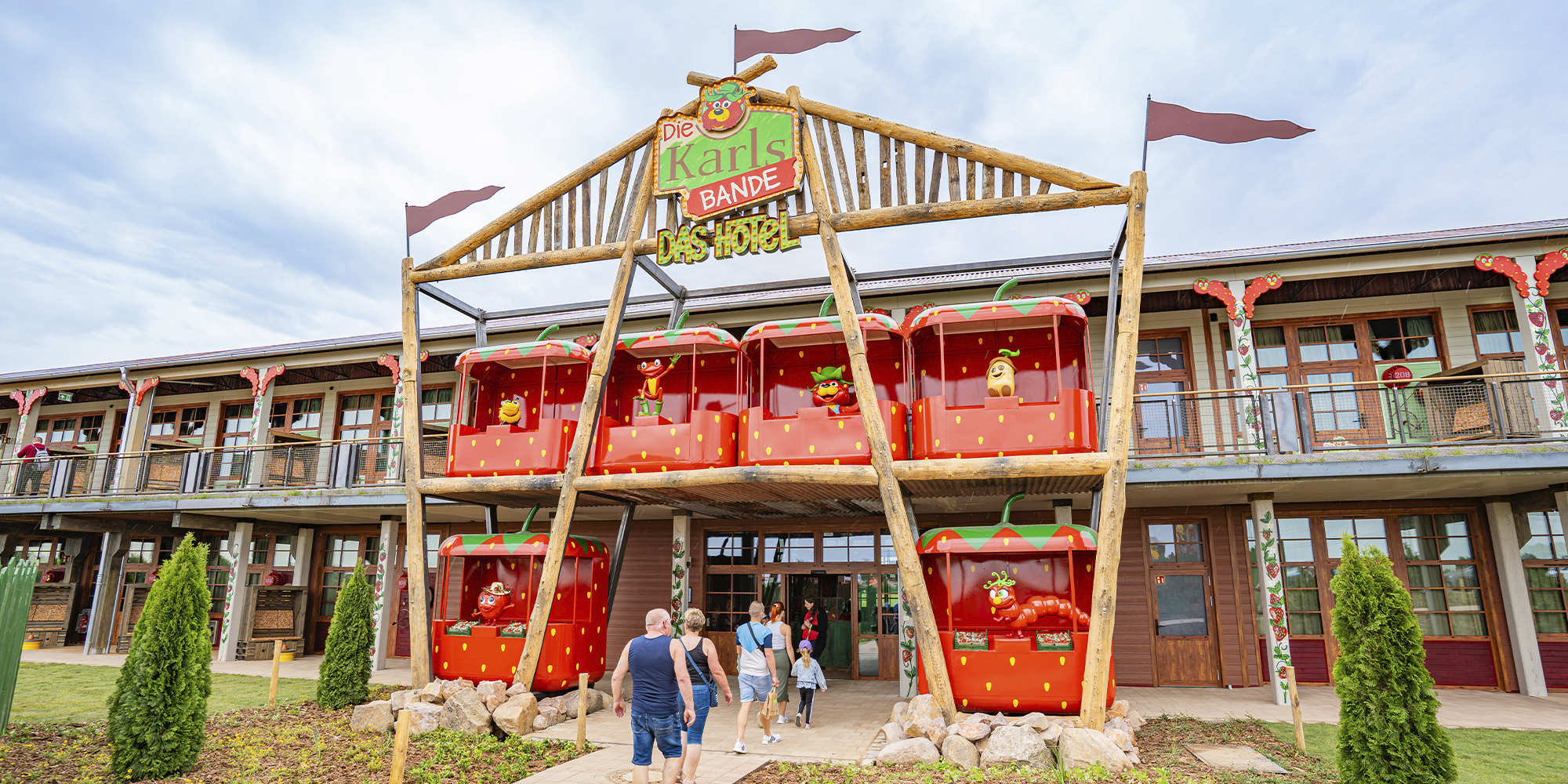 Außenansicht des Hotels „Die Karls Bande“ in einem Karls Erlebnis-Dorf. Der zweistöckige Holzbau ist mit einer markanten Fassade gestaltet, in die sechs riesige, rote Erdbeergondeln integriert sind. In den Gondeln befinden sich verschiedene bunte Maskottchen-Figuren. Vor dem Haupteingang spaziert eine Gruppe von Gästen über den gepflasterten Vorplatz. Über dem Eingang prangt ein rustikales Holzschild mit dem Logo „Die Karls Bande – Das Hotel“.