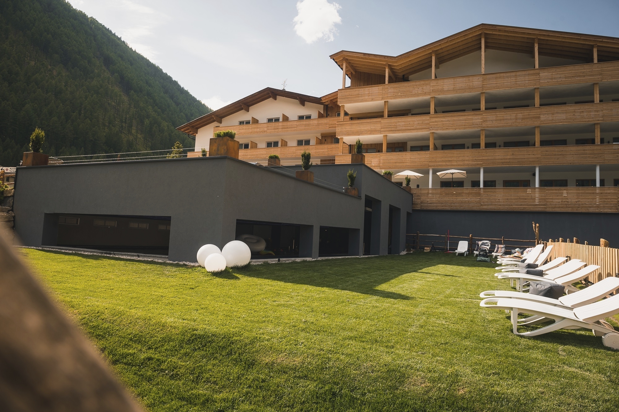 Moderne Hotelanlage in den Bergen mit großzügigen Holzfassaden und Balkonen. Im Vordergrund befindet sich eine gepflegte grüne Liegewiese mit weißen Sonnenliegen und dekorativen Leuchtkugeln. Das Gebäude schmiegt sich an einen bewaldeten Berghang unter sonnigem Himmel.