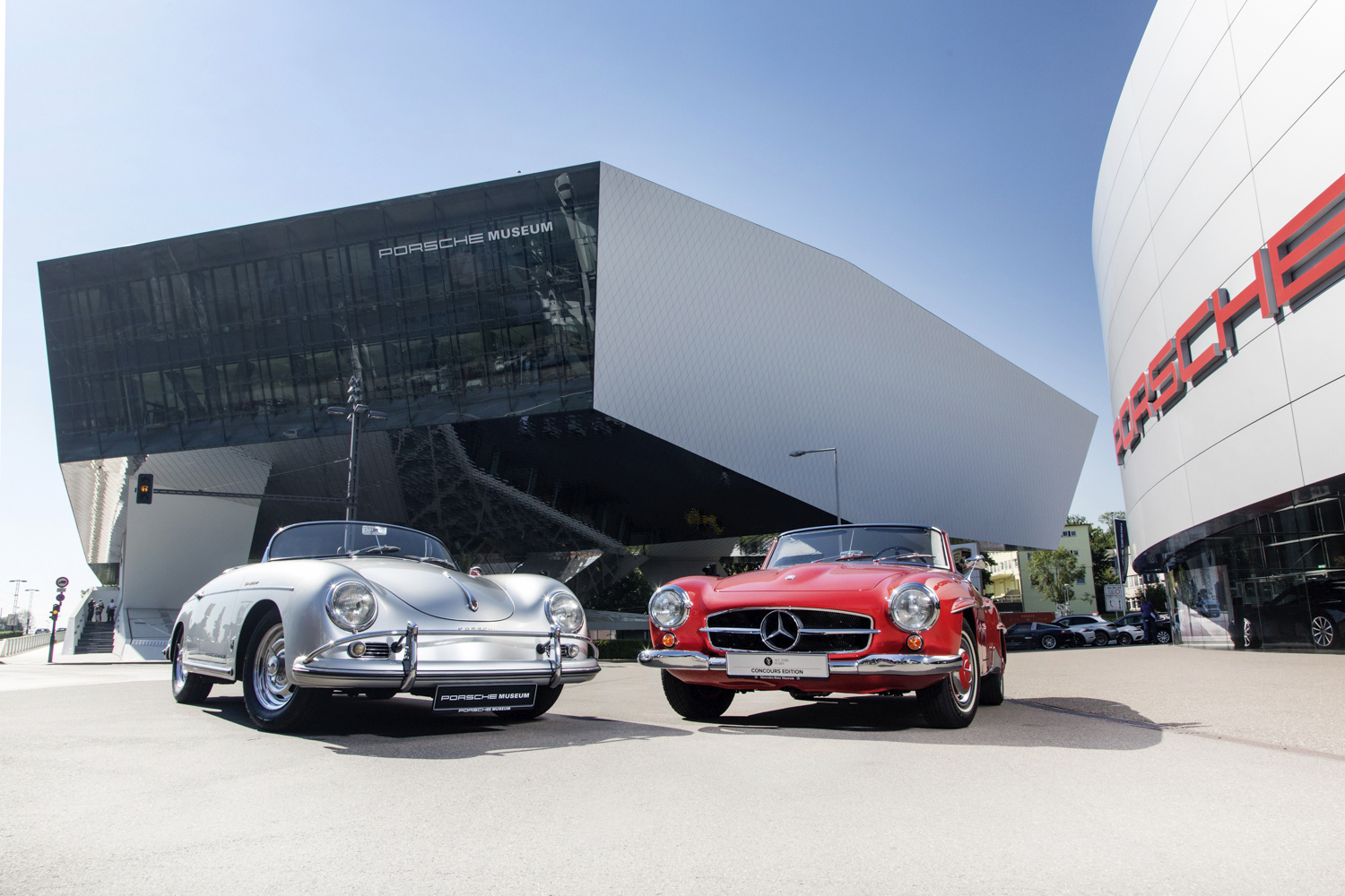Ein silberner Porsche-Klassiker und ein roter Mercedes-Benz Oldtimer stehen nebeneinander auf dem Vorplatz des Porsche Museums in Stuttgart. Im Hintergrund ist die markante, futuristische und kantige Architektur des Museumsgebäudes unter blauem Himmel zu sehen.