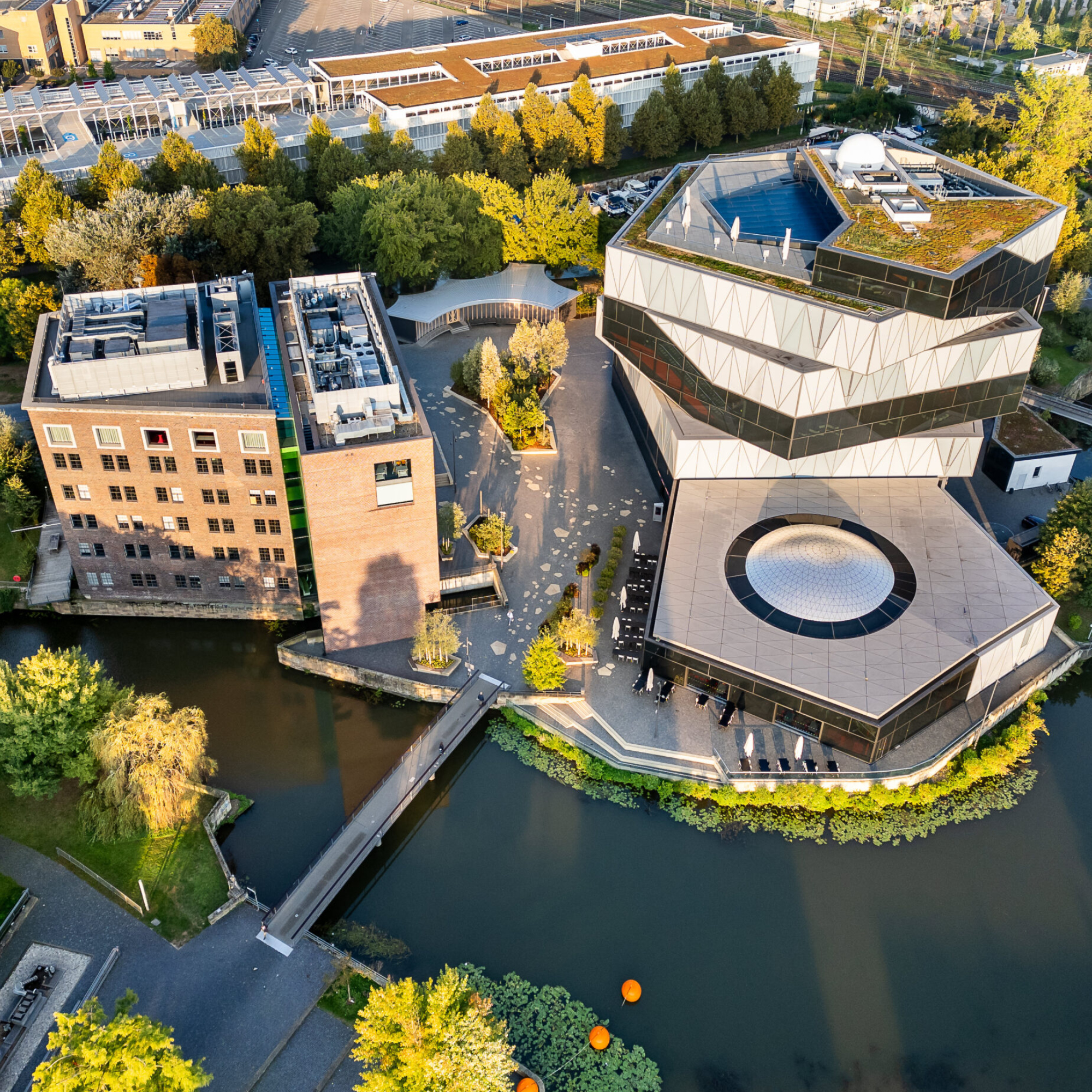 Luftbild des experimenta Science Centers in Heilbronn, direkt am Neckar gelegen. Zu sehen ist der markante Neubau mit seiner gläsernen, spiralförmigen Architektur und begrüntem Dach sowie der angrenzende Backsteinbau des Bestandsgebäudes. Eine Brücke führt über das Wasser zum Vorplatz der Anlage, die von Bäumen und Parkflächen umgeben ist.
