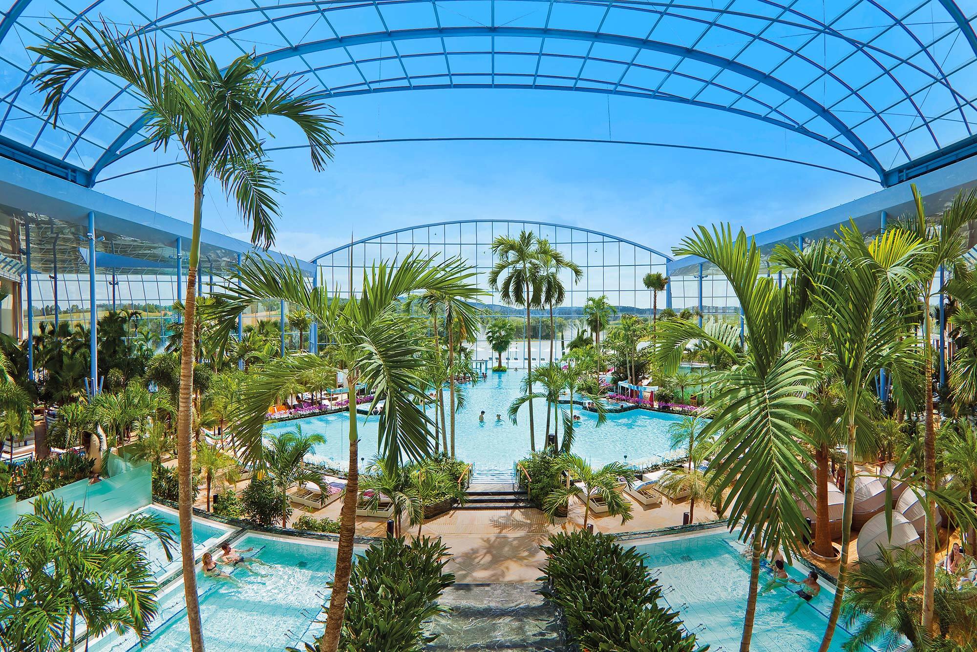 Blick von oben auf eine lichtdurchflutete Thermenlandschaft mit Palmen und Pool unter einer Glaskuppel.