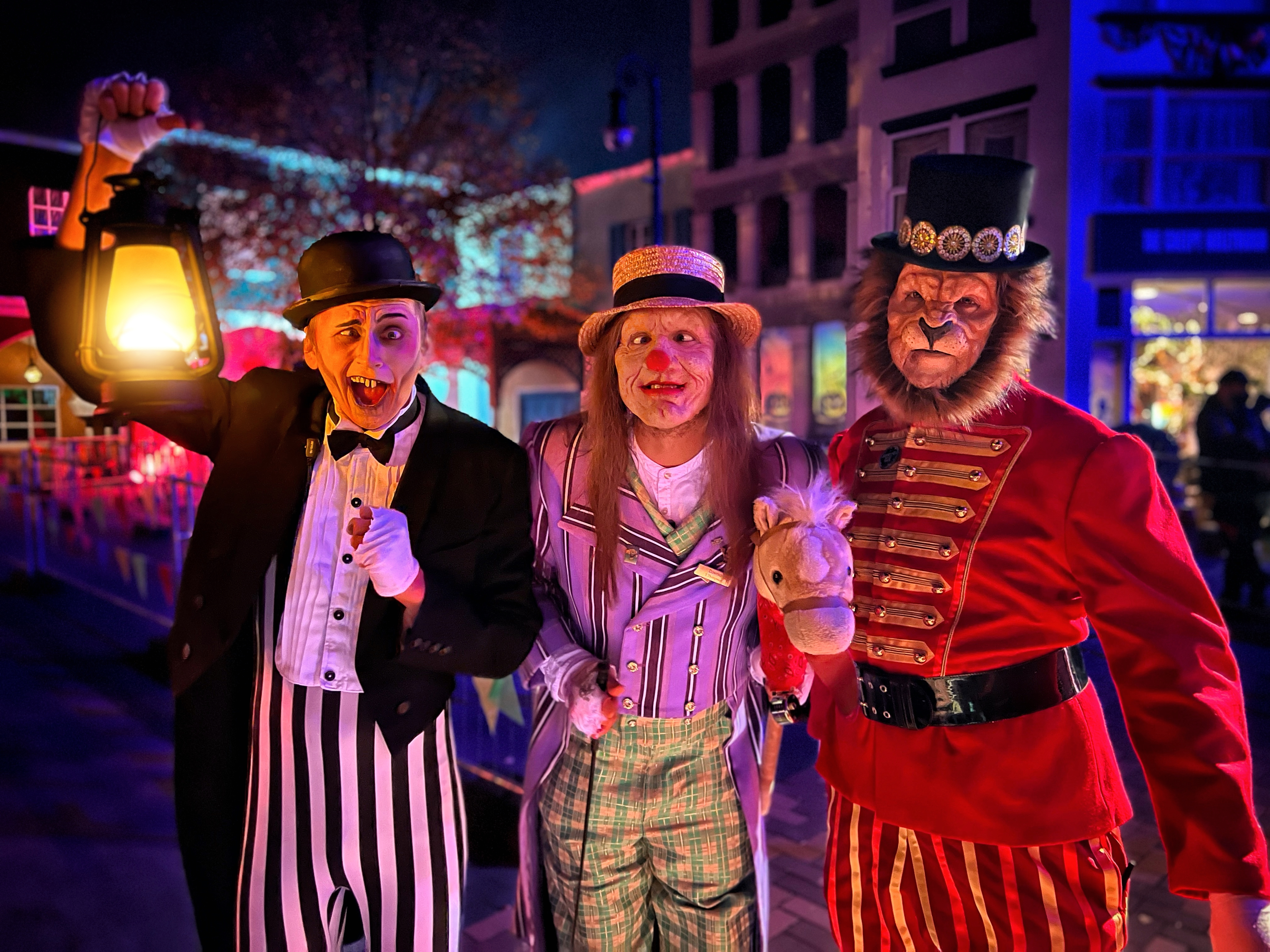 Drei schaurig verkleidete Darsteller in bunter Zirkus- und Zylinderkleidung posieren bei Nacht.