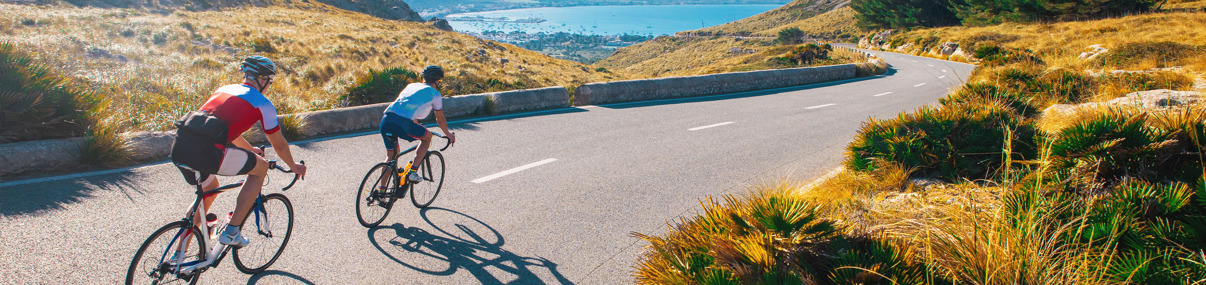 Zwei Rennradfahrer auf einer langen Straße und im Hintergrund das blaue Meer.