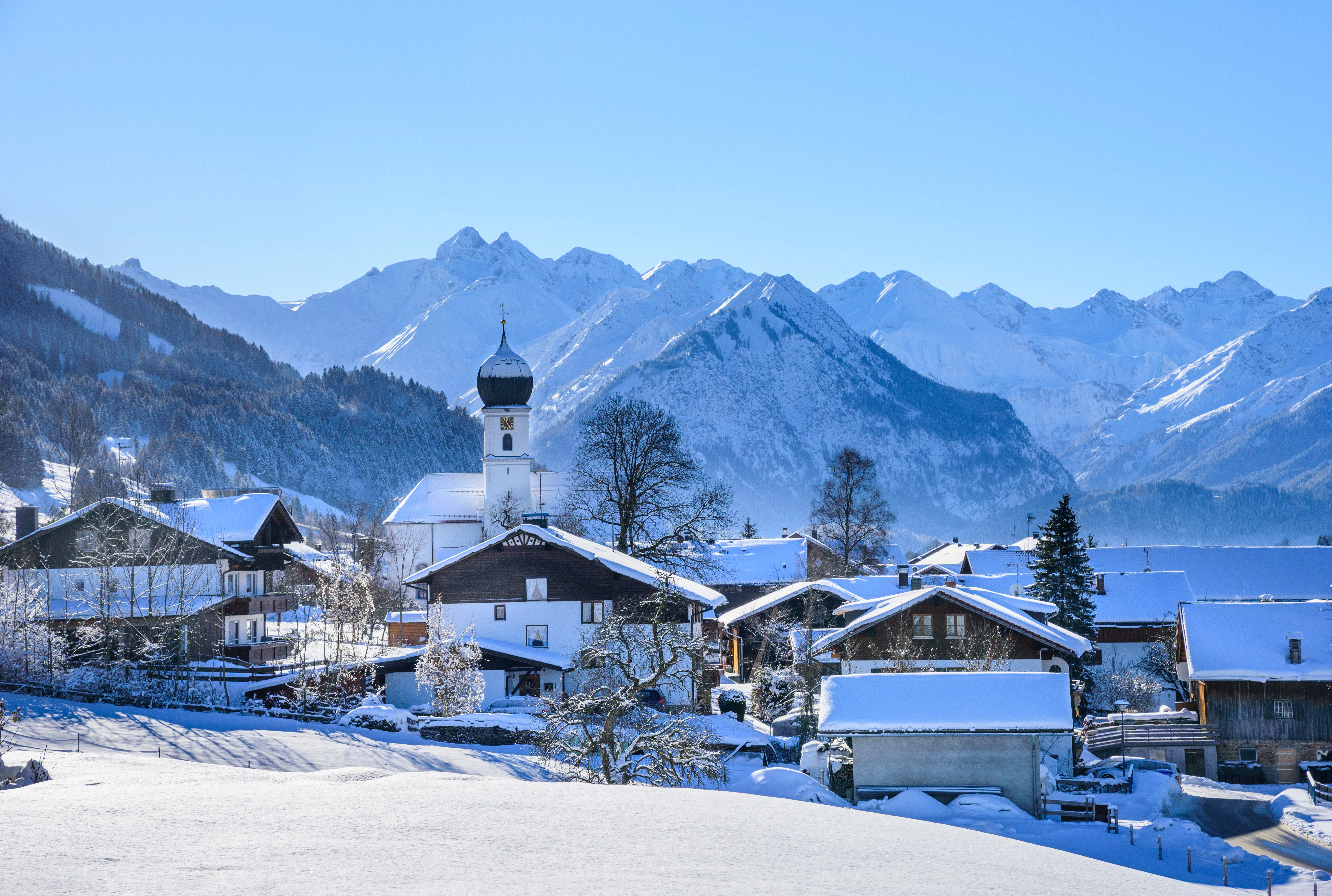 Verschneites Bergdorf mit Zwiebelturmkirche vor imposantem Alpenpanorama.