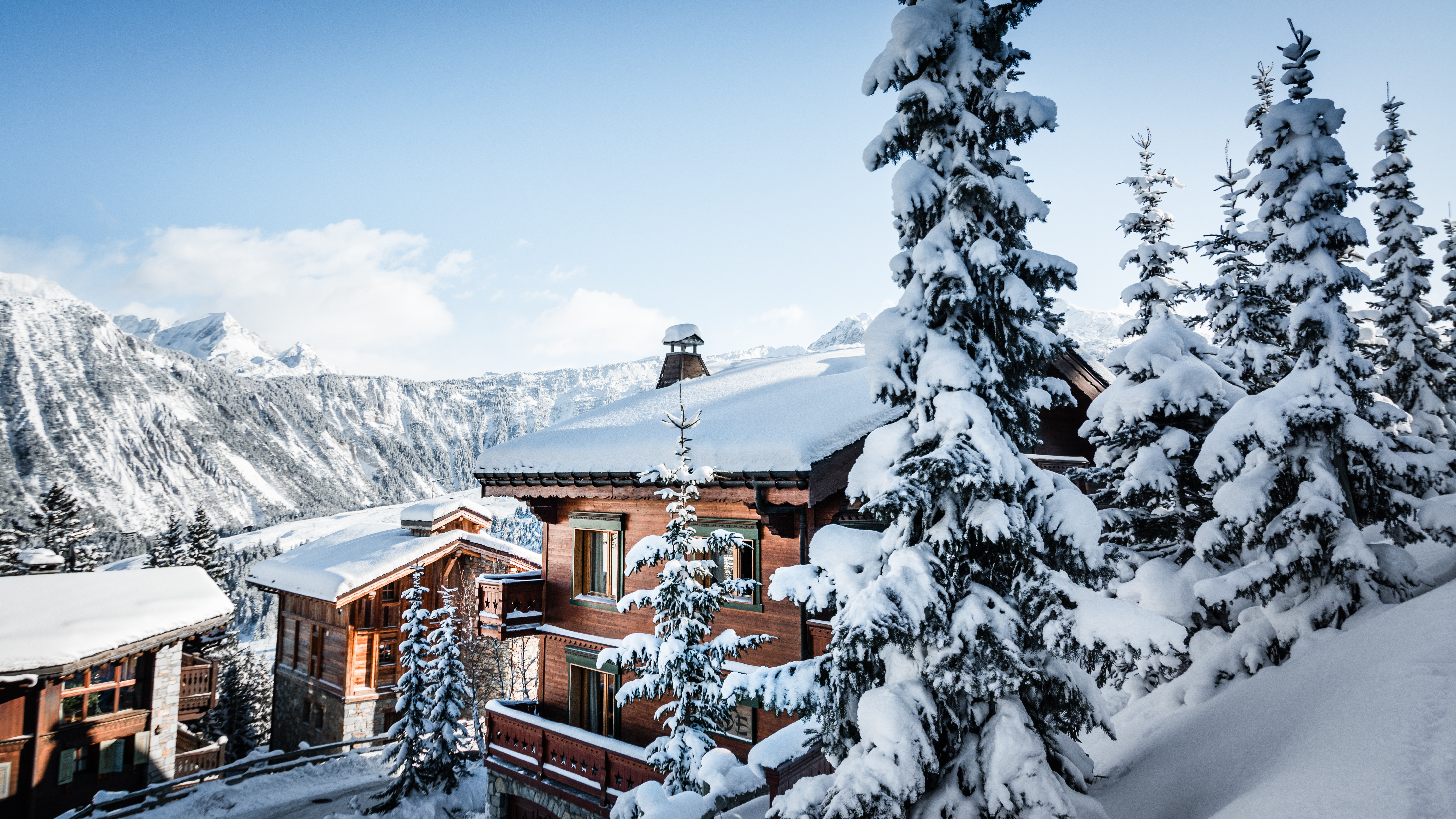 Verschneite Holzchalets an einem Berghang neben tief verschneiten Tannen.