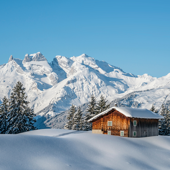Einsame Holzhütte auf einer tief verschneiten Wiese vor schneebedeckter Bergkette.