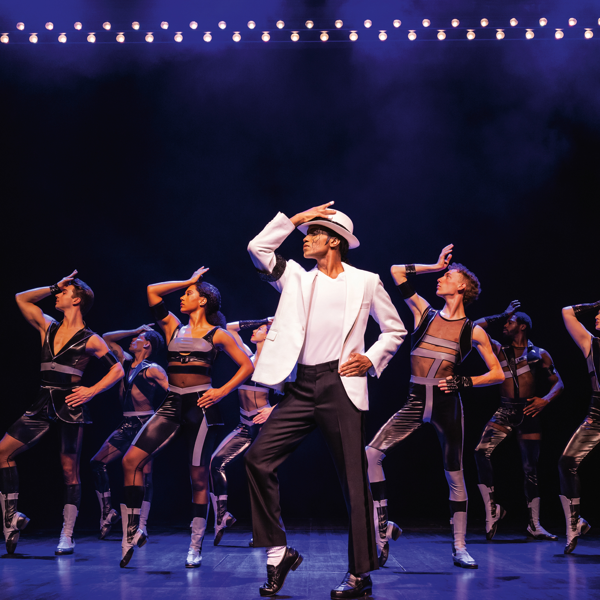Eine spektakuläre Bühnenszene aus dem Michael Jackson Musical. Im Zentrum steht der Hauptdarsteller in ikonischer Pose mit weißem Sakko, schwarzer Hose und Hut, die Arme triumphierend erhoben. Er wird von sechs Tänzern in schwarzen, glitzernden Kostümen umgeben, die in verschiedenen choreografierten Positionen auf der Bühne knien. Der Hintergrund zeigt eine stilisierte Skyline und zahlreiche leuchtende Neonreklamen, die eine nächtliche Großstadtatmosphäre erzeugen.
