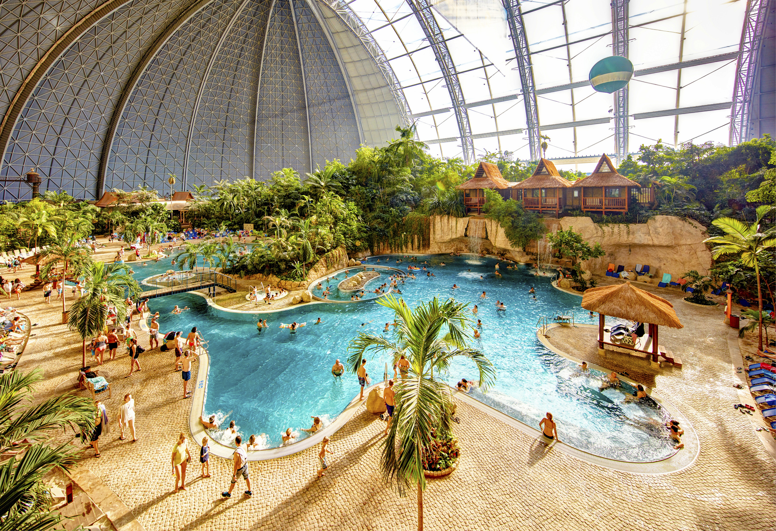 Blick in die riesige Tropenhalle des Tropical Islands mit einer weitläufigen, hellblauen Lagunen- und Poollandschaft. Zahlreiche Badegäste entspannen im Wasser und auf den umliegenden Wegen, die von echten Palmen und üppigen, grünen Pflanzen gesäumt sind. Im Hintergrund sind strohgedeckte Bungalows auf einer künstlichen Felswand mit Wasserfällen unter der gewaltigen Kuppelkonstruktion zu sehen.
