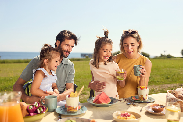 Familie sitzt fröhlich im Freien an einem gedeckten Esstisch mit Früchten und Getränken.