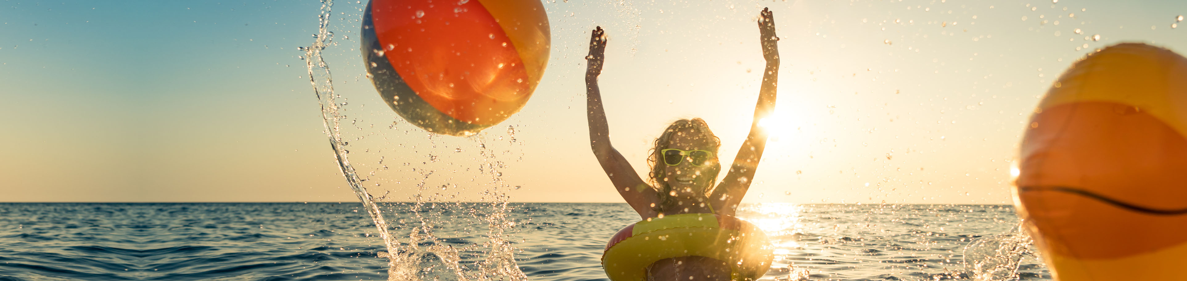 Kind planscht mit Schwimmreifen im Meer und wirft einen Wasserball hoch. Im Hintergrund ist ein Sonnenuntergang zu sehen. 