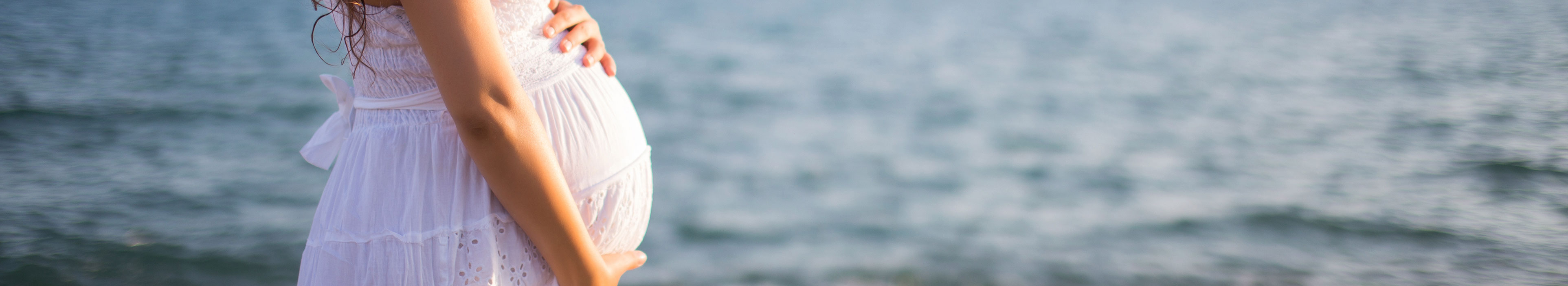 Schwangere Frau in einem weißem Kleid blickt auf das Meer