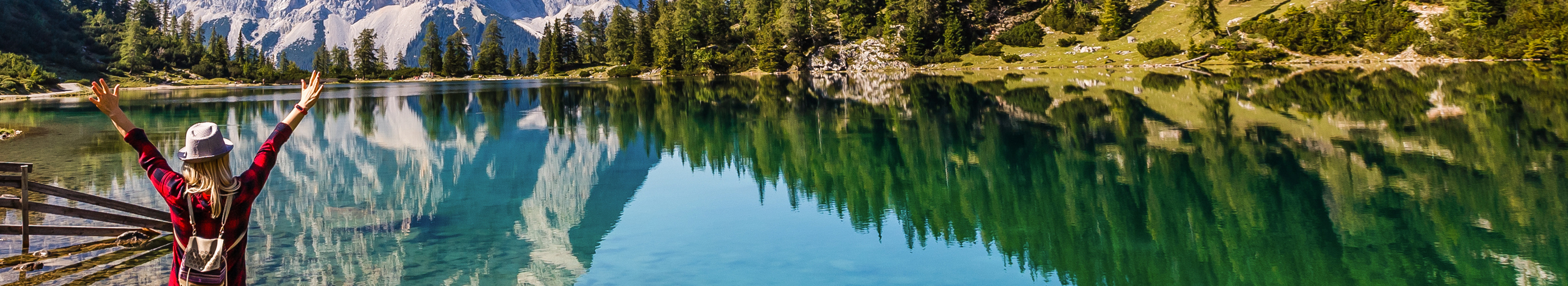Eine Reisende in warmer Kleidung von hinten, die an der Küste eines Bergsees steht und den Blick auf die Alpen in Österreich genießt