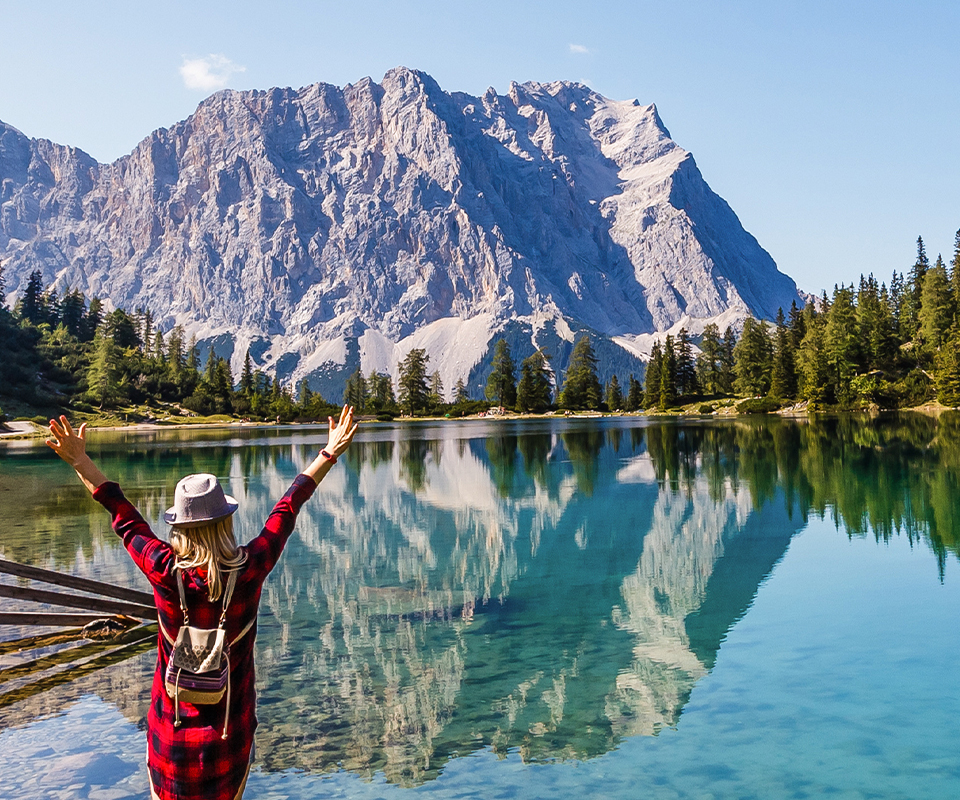 Eine Reisende in warmer Kleidung von hinten, die an der Küste eines Bergsees steht und den Blick auf die Alpen in Österreich genießt