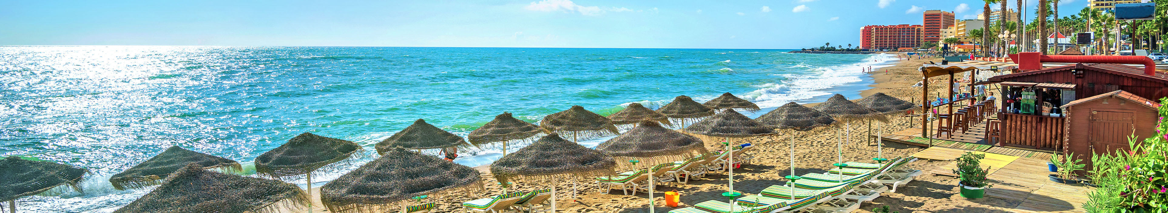 Strand in Spanien mit Sonnenschirmen und Liegen, Palmen und Hotels im Hintergrund. 