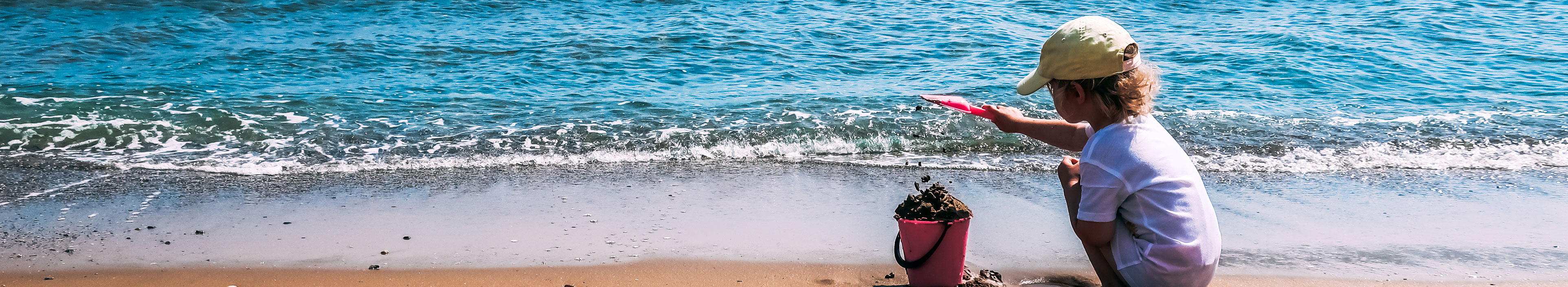 Ein Kind spielt mit einem rosa Plastikeimer und einer Schaufel im Sand an einem Strand in der Nähe des Meeres. Kreta, Griechenland.