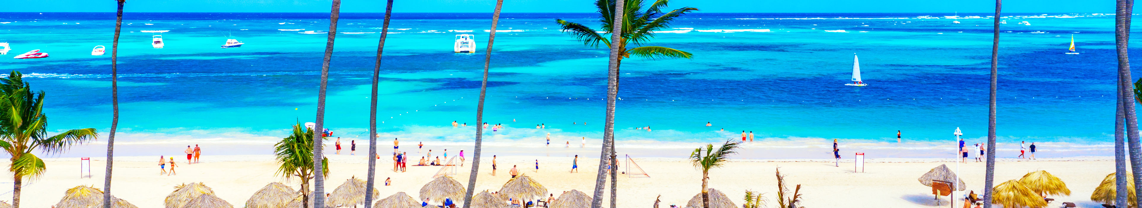 Strand in der Dominikanischen Republik mit weißem Sand, Palmen, blauem Meer und einigen Booten. Touristen schwimmen im Meer oder laufen am Strand.