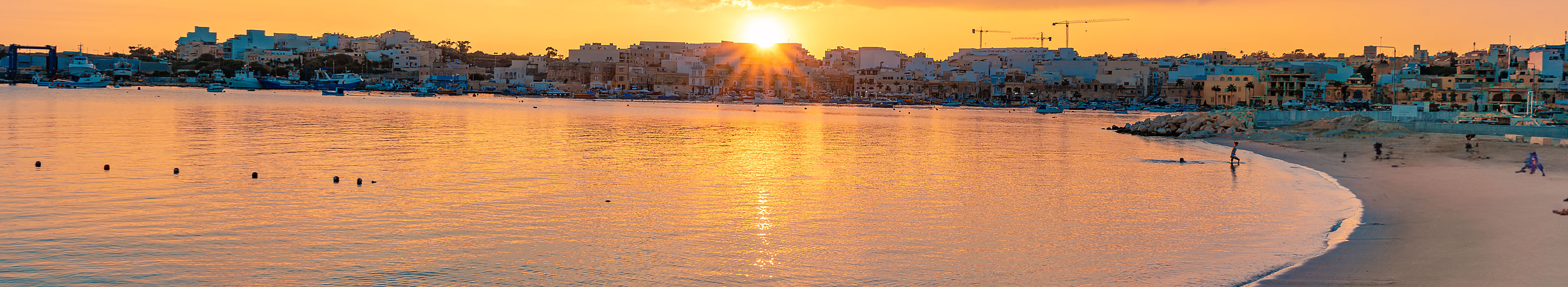 Strand auf Malta während eines Sonnenuntergangs, im Vordergrund blaue Sonnenschirme.