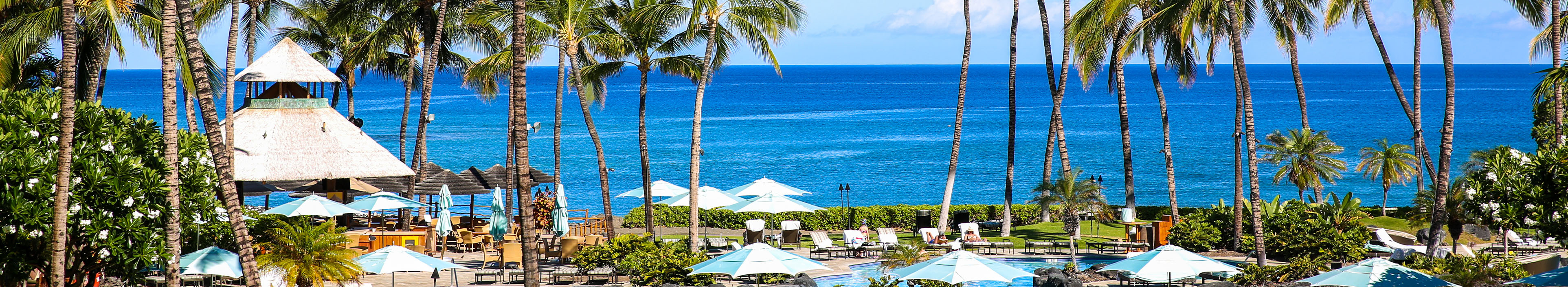 Palmen, ein Hoteltool, Sonnenschirme, und Liegen auf Hawaii, im Hintergrund das Meer.