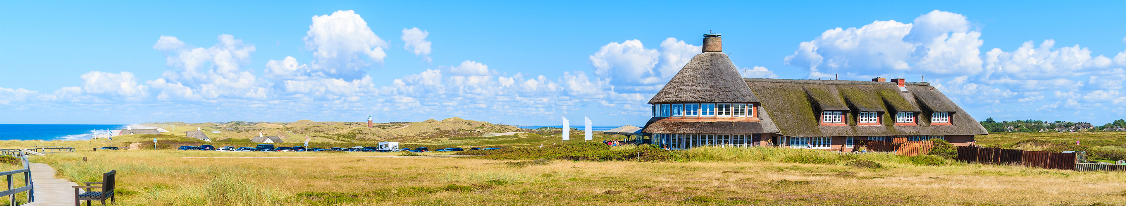  Bank am Fußweg entlang der Küste der Insel Sylt und typisch friesisches Gasthaus im Hintergrund.