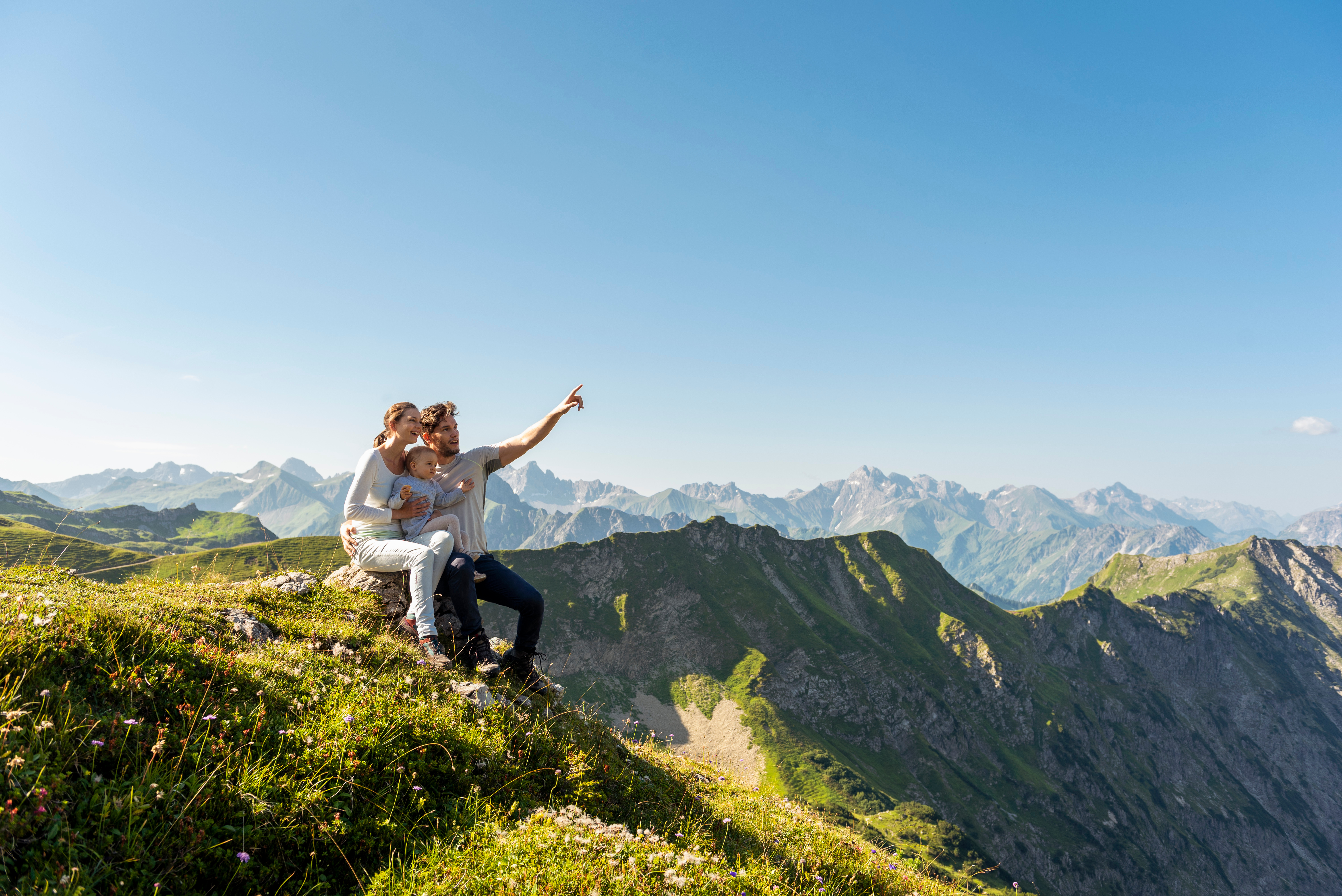 Ein Paar sitzt mit einem Kleinkind auf einem Felsen auf einem grasbewachsenen Berggipfel...