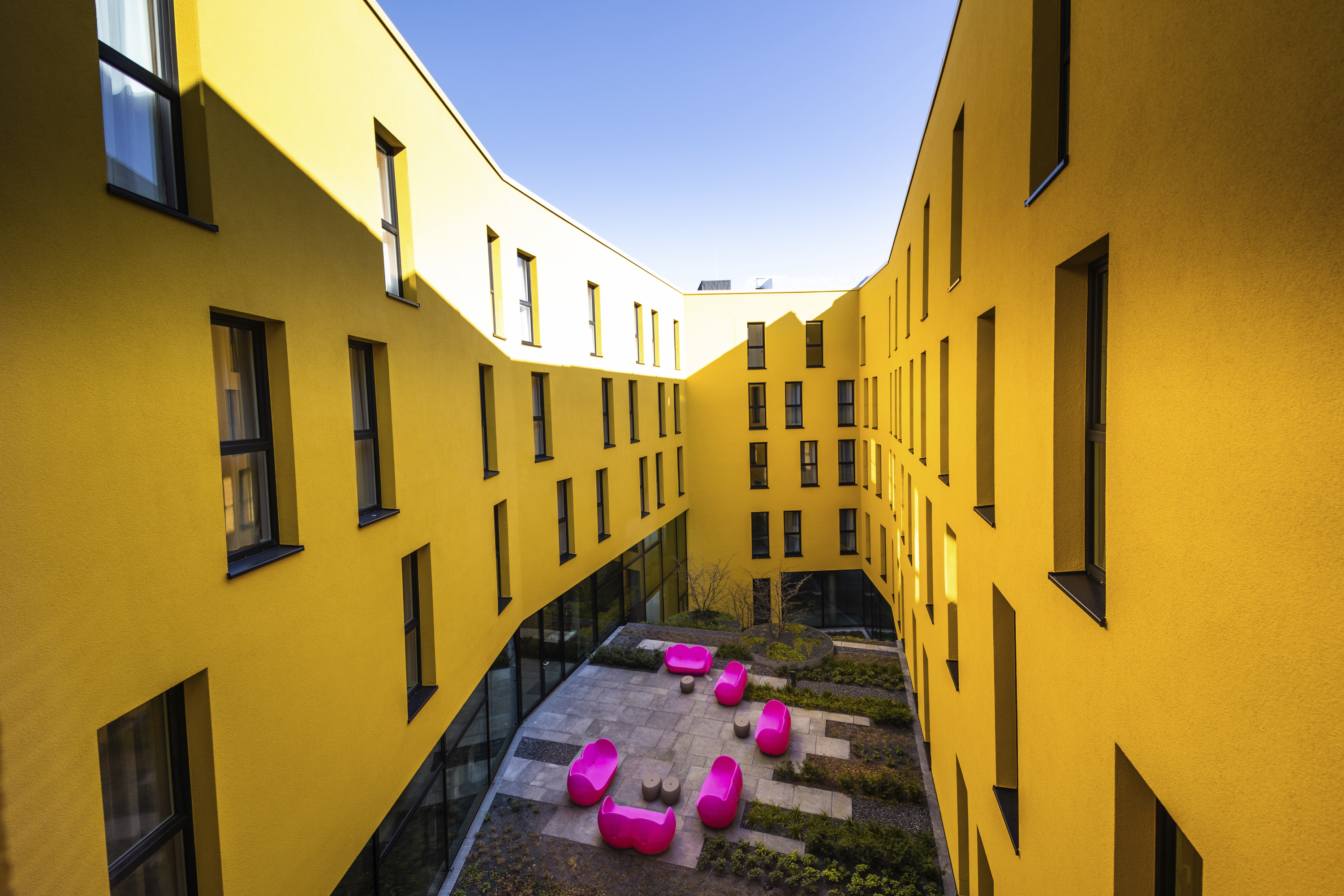 Innenhof mit gelben Gebaeudewaenden und pinken Sitzmoebeln.
